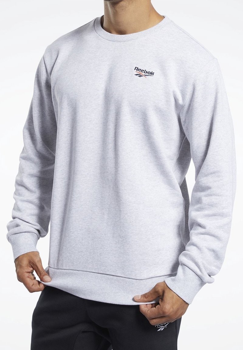 Szara bluza z okrągłym dekoltem, wykonana z miękkiego materiału, z małym czerwono-niebieskim logo Reebok na piersi oraz ściągaczami na mankietach i u dołu.