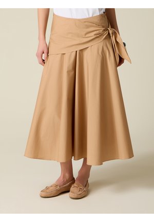 Femme portant une jupe midi beige taille haute avec un détail de nœud sur le côté et des mocassins assortis à enfiler.