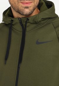 Sudadera Nike de color verde oliva con cremallera frontal, cordones negros y un sutil logo negro en el pecho. Tejido texturizado, corte atlético.