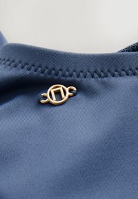 Blauwe stof met fijne ribbelstructuur en een gouden circulaire accent met een geometrisch patroon, gestikt langs de rand.
