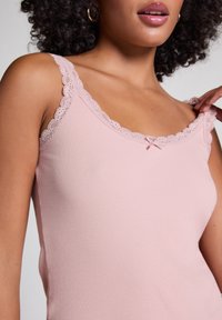 Débardeur côtelé rose clair avec une bordure en dentelle le long de l'encolure et des bretelles, agrémenté d'un petit nœud au centre.