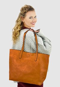 Grote oranje leren tote bag met twee handvatten aan de bovenkant, een stevige textuur en een minimalistisch design. Draagt over de schouder tegen een grijze achtergrond.