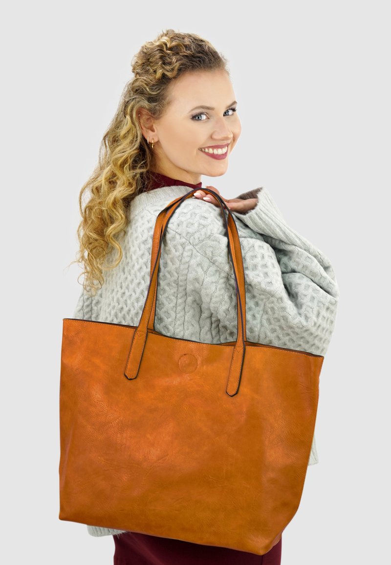 Grote oranje leren tote bag met twee handvatten aan de bovenkant, een stevige textuur en een minimalistisch design. Draagt over de schouder tegen een grijze achtergrond.