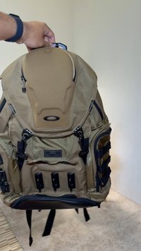 Ręka trzyma beżowo-czarny plecak Oakley z wieloma przegródkami i czarnymi paskami na tle gładkiej beżowej ściany.