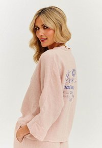 Svetlo ružová ľanová blúzka s dlhými rukávmi, voľný strih a okrúhly výstrih. Na zadnej strane je modrý potlačený text a dizajn slnka.