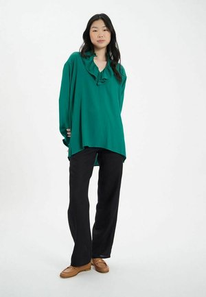 Blouse bleu-vert avec un col volanté, des manches longues et une coupe ample, associée à un pantalon large noir et des mocassins beige clair.
