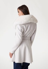 Manteau blanc avec un large col en fausse fourrure, taille ceinturée, ourlet évasé et manches longues, présentant une texture douce et une coupe structurée.