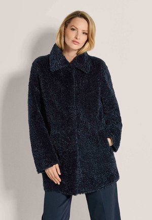 Blonde vrouw met een donkerblauwe, pluizige jas met kraag, staand tegen een lichte achtergrond met één hand in de zak.
