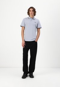 Lys grå polo shirt med krave, sort detaljering og logo, parret med sorte bukser og sorte sko, sat mod en ensfarvet hvid baggrund.
