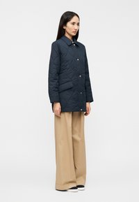 Μπλε ναυτικό μπουφάν με επένδυση, γιακά, χρυσά εξαρτήματα και πλαϊνή τσέπη, σε συνδυασμό με ανοιχτό μπεζ παντελόνια με φαρδιά πόδια και μαύρα παπούτσια.