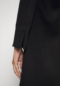 Prenda de lana negra de manga larga con un puño ligeramente acampanado, textura suave y corte holgado, mostrando una mano descansando sobre la tela.