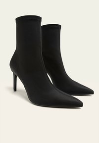 Mustat nilkkapituisten saappaat, jotka on valmistettu sileästä kankaasta, jossa on teräväkärkiset kärjet ja ohuet stiletto-korkot, ja näkyvät ompelusaumat saumoissa.