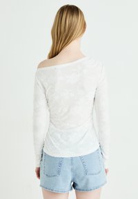 Hvit langermet topp laget av teksturert stoff med blomster mønstre, med en av-skulder design. Kombinert med lyseblå denimshorts.