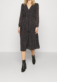 Vrouw in een zwarte midi-jurk met een klein beige patroon, lange mouwen, V-hals en zwarte hielenkorte laarzen.