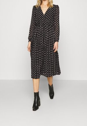 Vrouw in een zwarte midi-jurk met een klein beige patroon, lange mouwen, V-hals en zwarte hielenkorte laarzen.