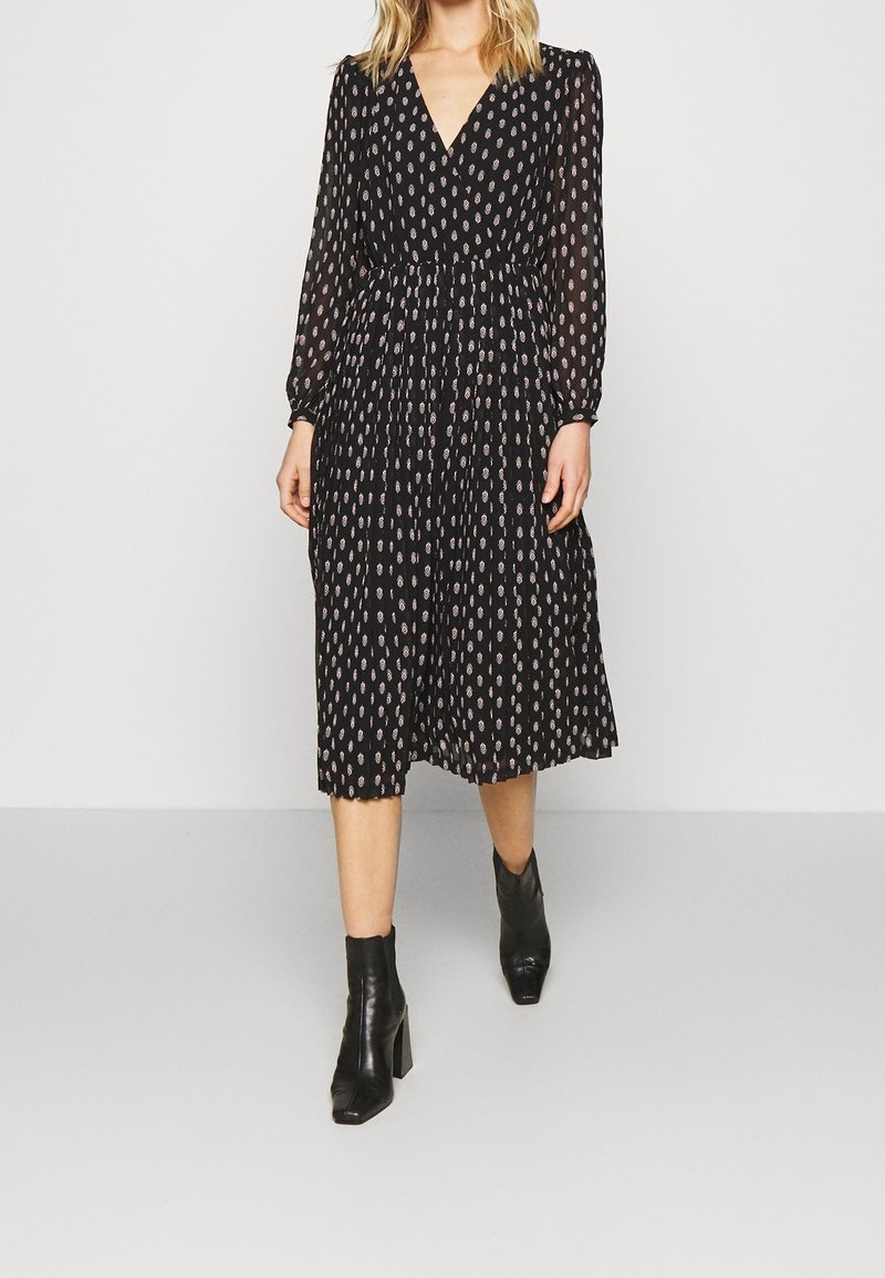 Vrouw in een zwarte midi-jurk met een klein beige patroon, lange mouwen, V-hals en zwarte hielenkorte laarzen.