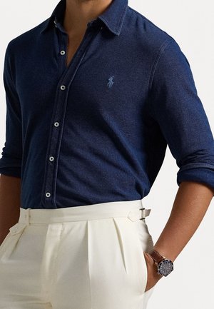 Homme portant une chemise à boutons bleu foncé avec les manches retroussées et un pantalon plissé taille haute crème, avec une montre à bracelet en cuir marron.