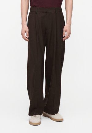 Homme portant un pantalon large marron foncé et des chaussures couleur crème, debout devant un fond clair uni.