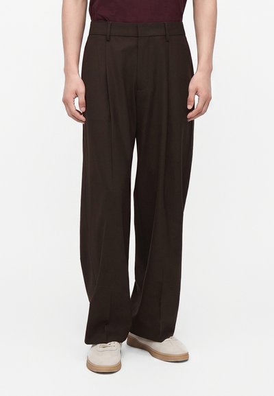 Homme portant un pantalon large marron foncé et des chaussures couleur crème, debout devant un fond clair uni.