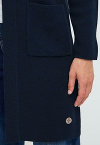 Cardigan en tricot marine foncé avec une poche à rabat et un accent de logo circulaire. Texture douce avec une coupe décontractée et un design ouvert à l'avant.