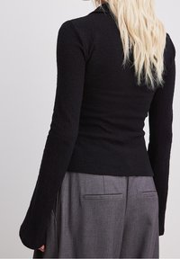 Schwarzes Langarmoberteil mit texturiertem Material und taillierter Passform. Kombiniert mit grauen, hochgeschnittenen Hosen mit Gesäßtaschen.