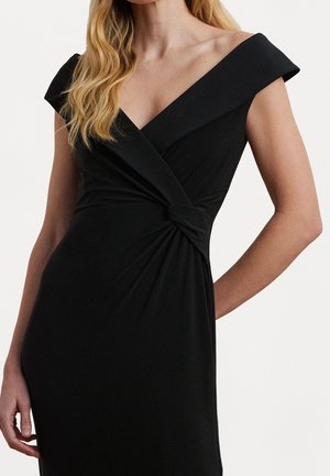 Vrouw in een zwarte, off-shoulder jurk met een geknoopt detail in de taille, lang blond haar, staand tegen een effen lichte achtergrond.