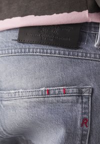 Jeans en denim bleu clair avec une étiquette en cuir noir. La couture rouge sur la poche arrière arbore un design en double 'R'. Texture douce, style décontracté.
