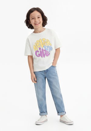 Gglimlachend kind met schouderlang haar draagt witte sneakers, opgerolde blauwe spijkerbroek en een wit T-shirt met kleurrijke tekst "Never Give Up".