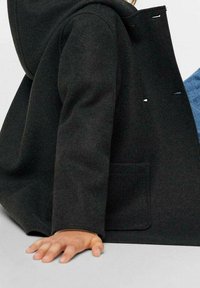 Svart ullkappa med huva, med en sidoficka och subtila sömdetaljer; ett barns hand vilar på en närliggande yta.