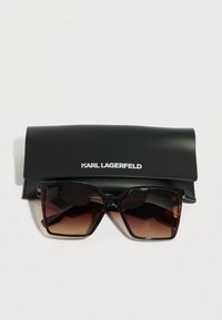 Gafas de sol grandes con degradado marrón y montura cuadrada, acompañadas de un estuche rígido negro con la marca.