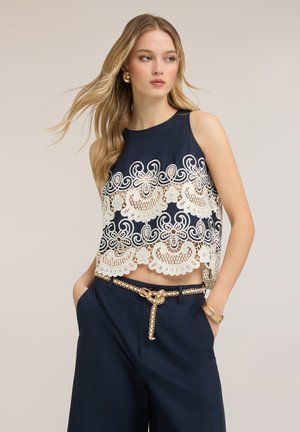 Mujer joven con top sin mangas azul marino con superposición de encaje floral blanco y pantalones azul marino, complementado con cinturón de cadena dorada y pulseras.
