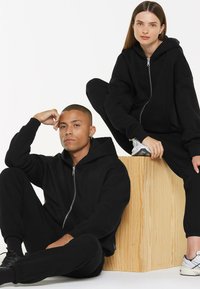 Black zip-up hoodie and matching pants made of soft material. Model poses seated on a wooden block, showcasing relaxed fit and style.