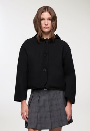 Giacca nera corta con cappuccio, dotata di grandi bottoni, una texture liscia e maniche ampie. Indossata sopra una gonna a pieghe grigia a fantasia tartan.