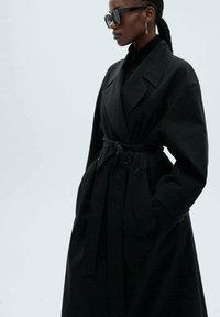 Schwarzer Trenchcoat aus strukturiertem Stoff, mit weitem Kragen, taillierter Taille und langen Ärmeln. Getragen mit schwarzen Sonnenbrillen.