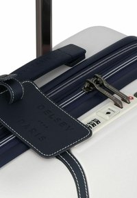 Maleta Delsey con una carcasa dura blanca, acentos en azul marino, una etiqueta de cuero, cremalleras dobles y un candado de combinación en el lateral.