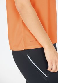 Orange snabbtorkande T-shirt med en mjuk textur, tillsammans med svarta leggings med vit sidrandedetalj. Närbild av arm och midja.