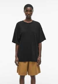 Camiseta negra de gran tamaño hecha de algodón con mangas amplias, que presenta un logo sutil. Combinada con pantalones cortos marrones hasta la rodilla.