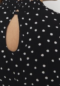 Svart tyg topp med vita prickar, med en smockad halsringning och en baköppning som säkras med två knappar.