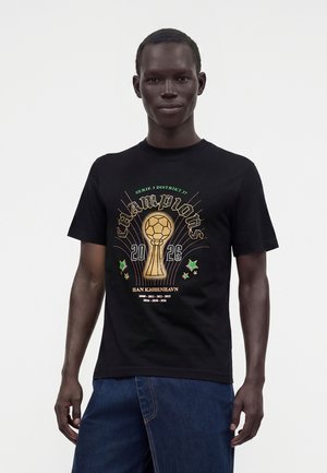 Ung mand iført en sort T-shirt med en gylden trofæ og teksten "champions 2026", kombineret med blå jeans mod en ensfarvet baggrund.