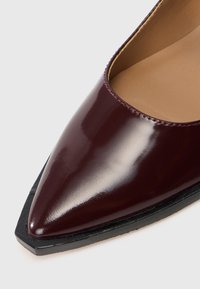 Zapato de charol burdeos con punta afilada, que presenta una textura suave y una suela negra. El detalle de borde cosido realza el diseño.