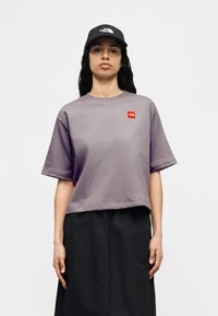 Γυναίκα που φοράει γκρι κοντομάνικο T-shirt με κόκκινο λογότυπο και μαύρο καπέλο με λευκό κείμενο, στέκεται μπροστά σε απλό ανοιχτό φόντο.