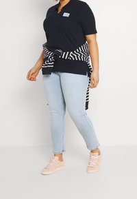 Personne portant un polo noir, un jean bleu clair, des baskets roses et un pull rayé noir et blanc noué autour de la taille.