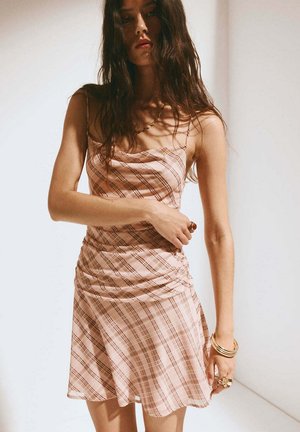 Mujer con cabello largo y oscuro, vistiendo un vestido rosa sin mangas con cuadros, pulseras y anillos dorados, de pie frente a un fondo claro y liso.