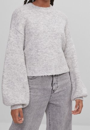 Personne portant un pull texturé gris clair à manches longues avec des manches volumineuses et un jean gris délavé taille haute.