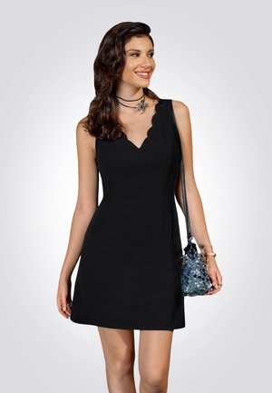 Vestito nero senza maniche con scollatura a onda, design aderente e texture del tessuto. Abbinato a una borsa a spalla scintillante e gioielli a strati.