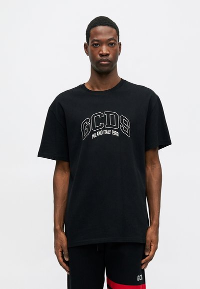 Juoda medvilnės marškinėliai su apykakle ir trumpomis rankovėmis. Priekyje yra baltas tekstas "GCDS MILANO ITALY 1998" drąsiu šriftu.