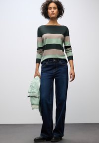 Gestreifter Pullover in Grün, Beige und Dunkelgrün, kombiniert mit dunkelblauen Schlagjeans. Hält eine mintgrüne Bomberjacke. Schwarze Stiefel.