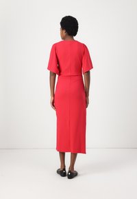 Abito midi rosso con maniche corte e larghe; vita aderente e gonna svasata; tessuto morbido; cucitura centrale sul retro; indossato con ballerine nere.