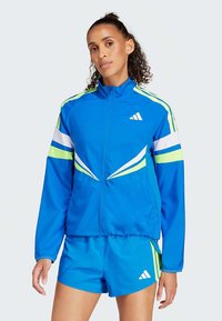 Blaue Sportjacke mit grünen und weißen Streifen, Reißverschluss und hohem Kragen. Kombiniert mit passenden blauen Shorts mit Logodruck.