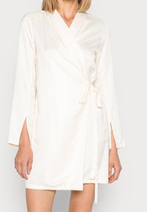 Femme portant une robe portefeuille longue en satin couleur crème, nouée à la taille avec des fentes latérales sur les manches, visible du cou aux cuisses.
